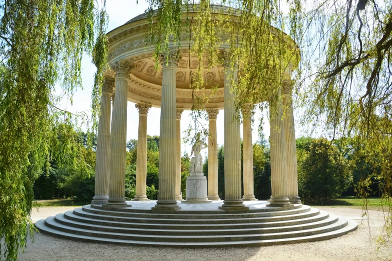 the fountain at the bottom of the tree filled yard has a column and pillars and the sculpture in the center is surrounded by columns