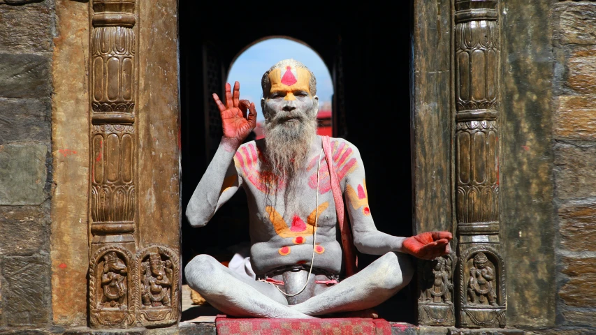 a seated god with painted face and long white beard is doing yoga