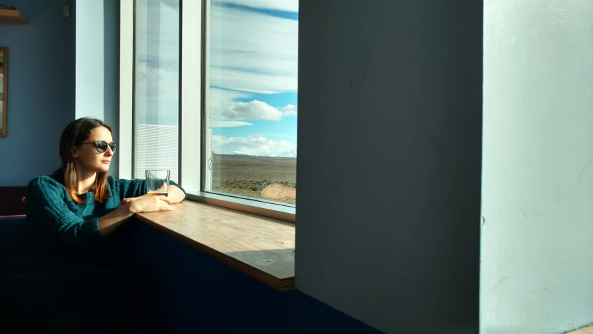 a woman wearing sunglasses leaning against a windowsill and looking out the window