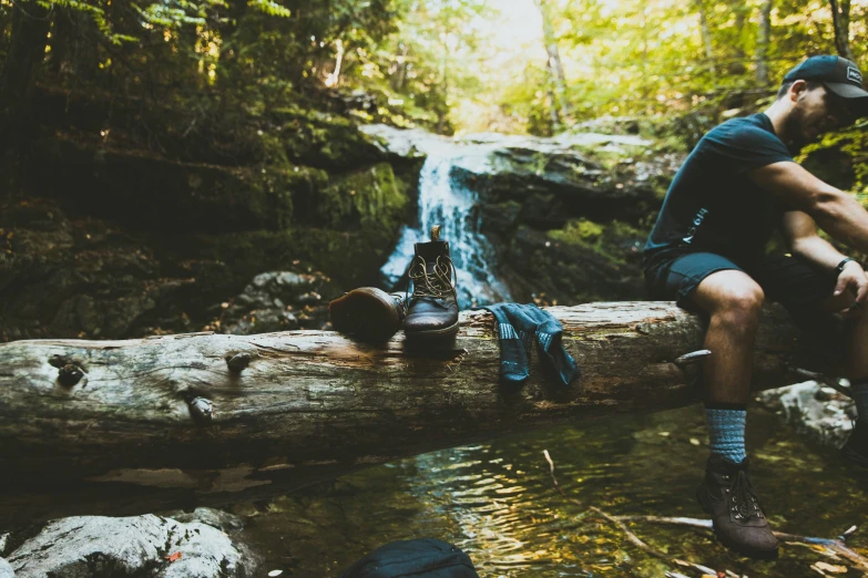 a man in shorts and boots sit on a log over a stream