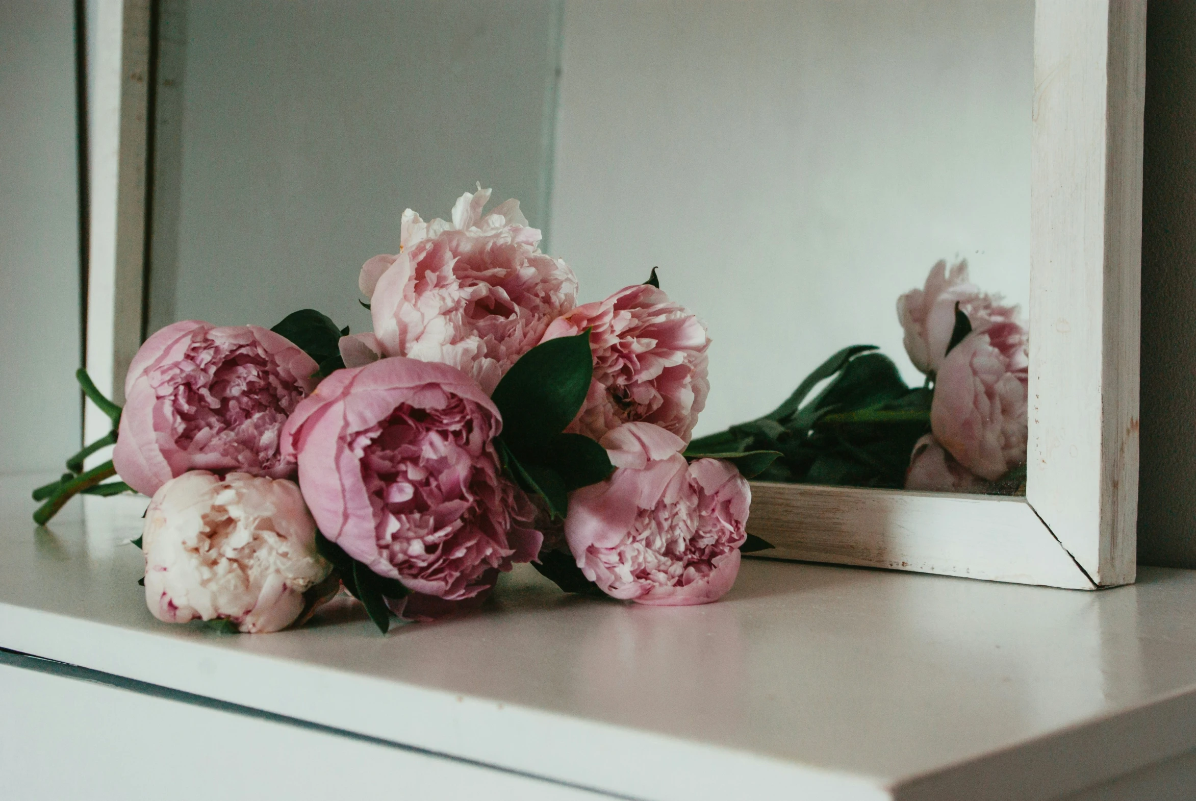 a white shelf topped with pink peonies in front of a mirror