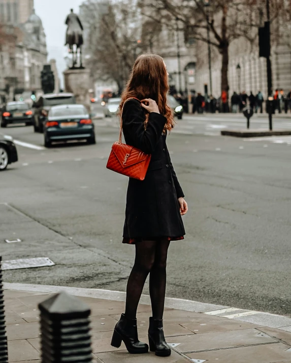 a woman standing on the side of a street wearing a black coat and a red purse