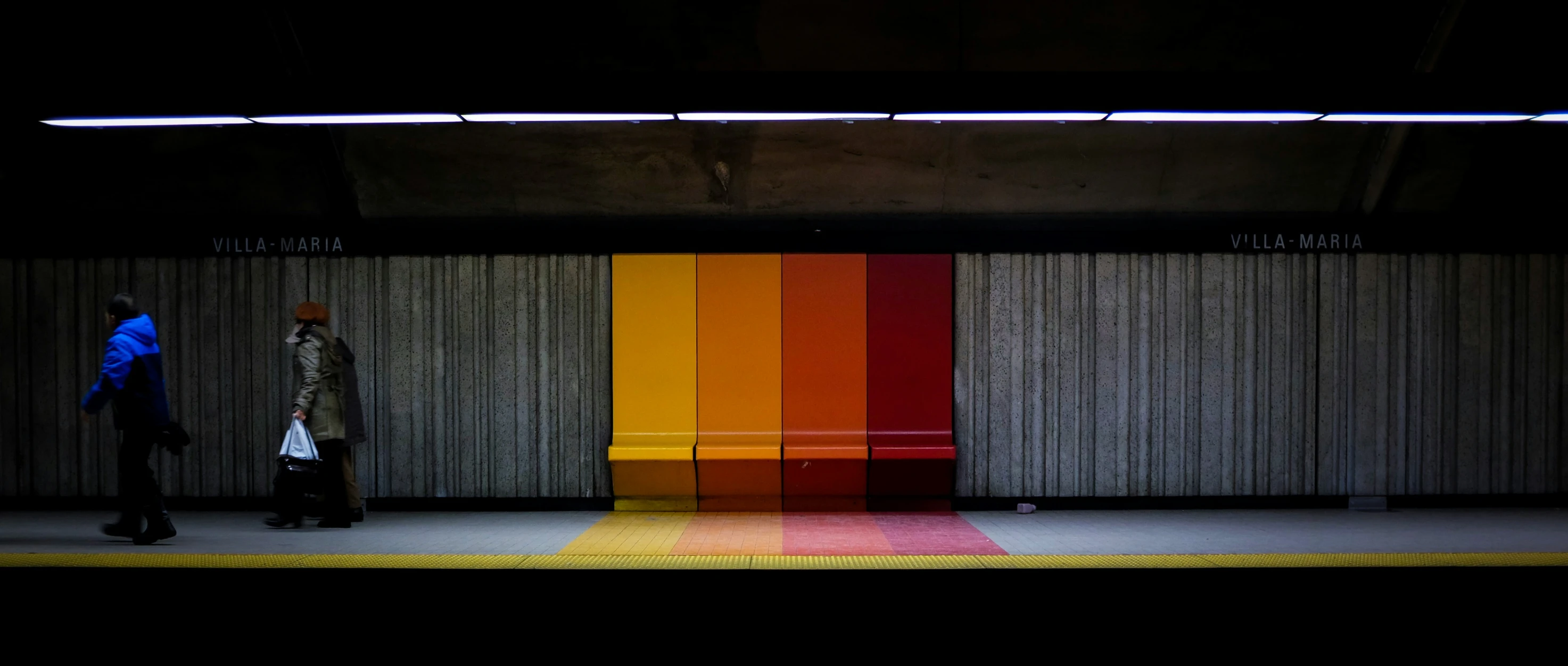 two people walking down a train platform, in front of a colored wall