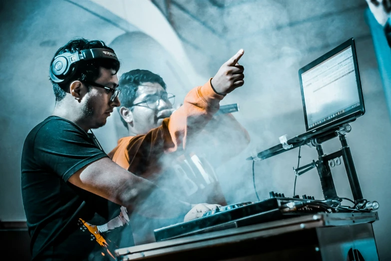 two men in hardhats and protective gear stand at a dj's turntable while another man looks on