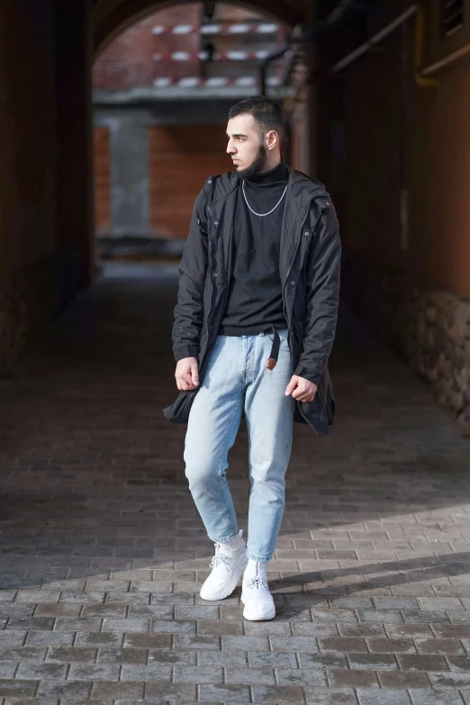 a man with short hair stands in an alley while wearing jeans and jacket