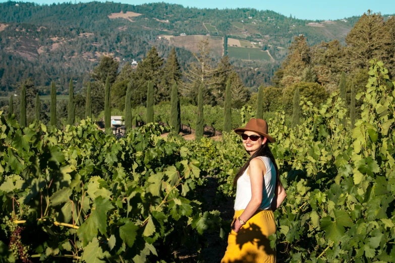 a woman in a hat and sunglasses is standing among the bushes