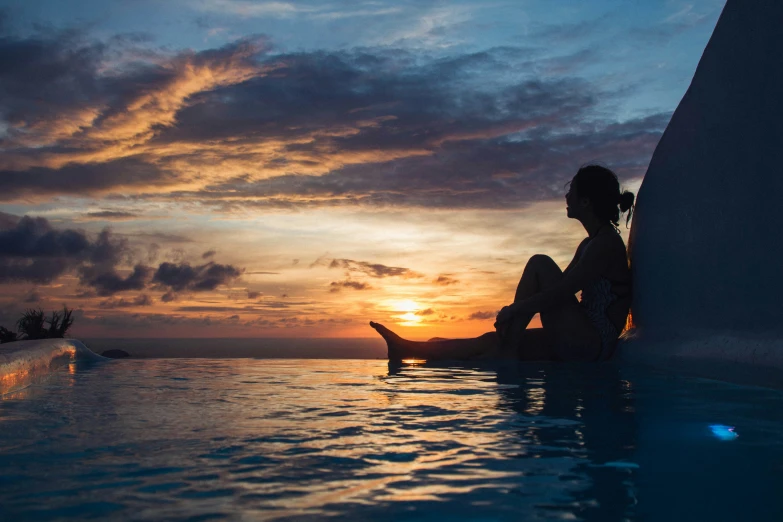 a woman is sitting on the edge of a pool in front of an ocean sunset
