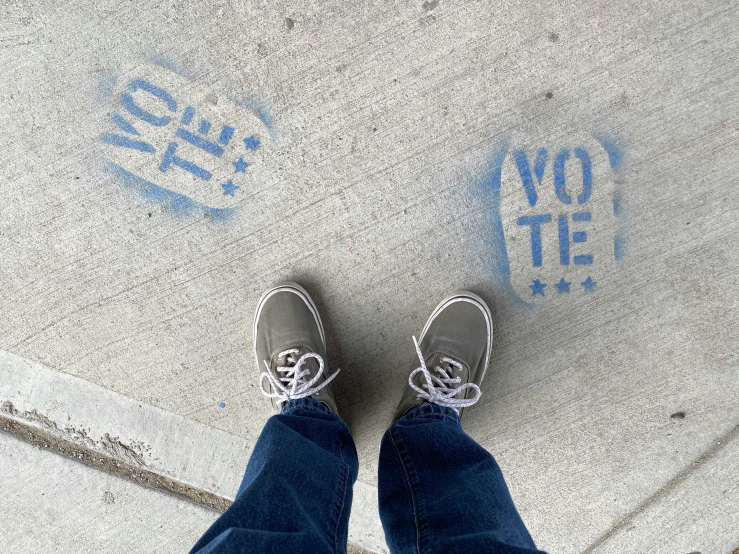 person's feet and shoes on an unpainted sidewalk with a vote stamp