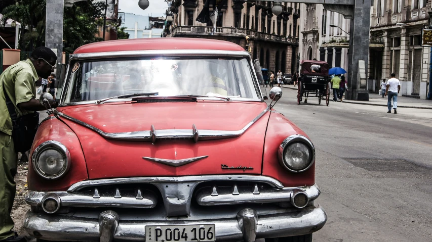 an old red car parked in front of a man on the sidewalk