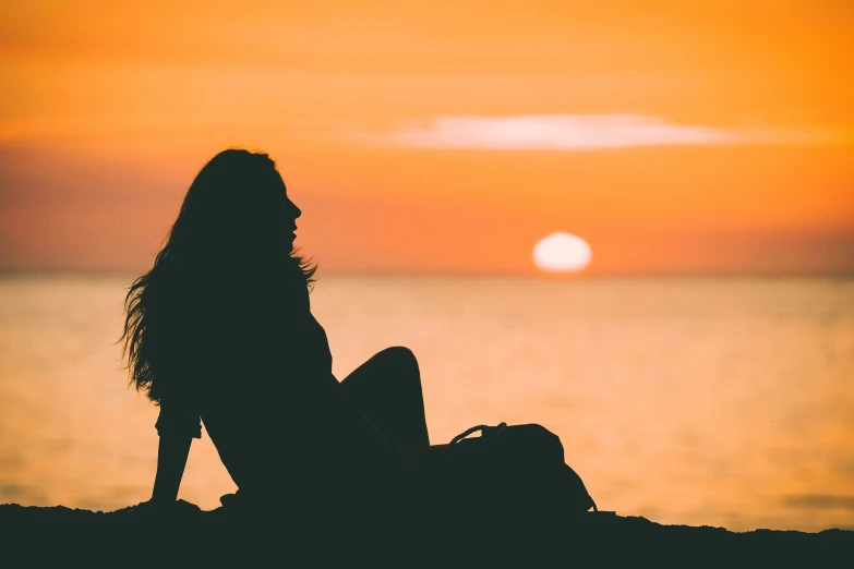 the silhouette of a girl sitting on a beach looking out at the ocean