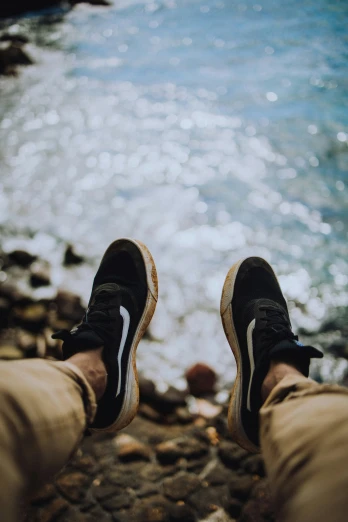 a person is wearing black sneakers with their feet on some rocks near the water