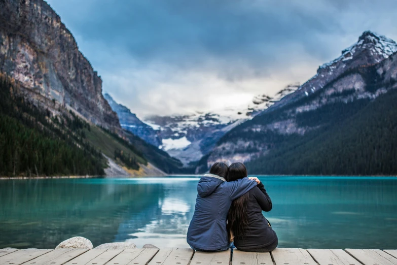 two people sit at the end of a dock overlooking a body of water with snow capped mountains in the distance