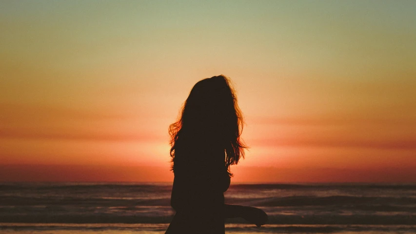 the back side of a girl standing near the water as the sun goes down