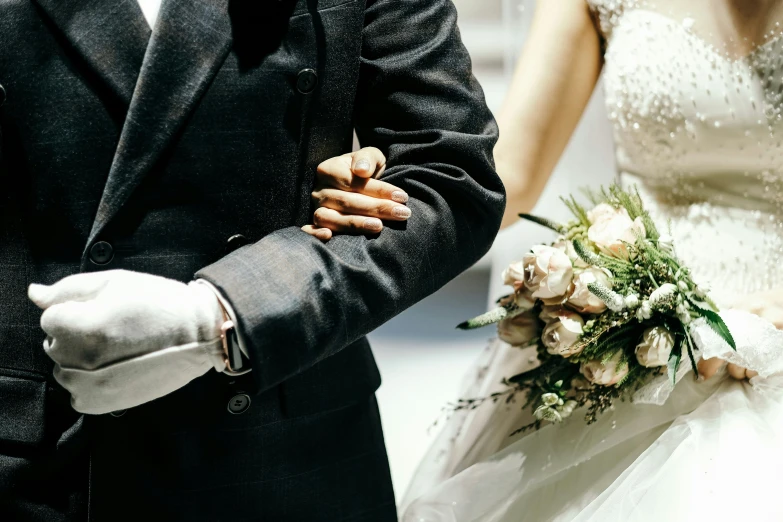 a bride in white gloves and dress is holding the groom's arm