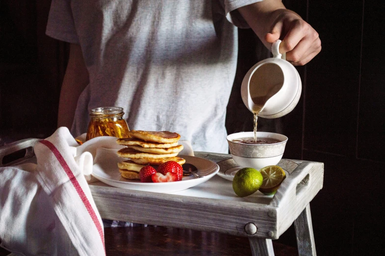 a person pouring syrup over pancakes and fruit on a tray