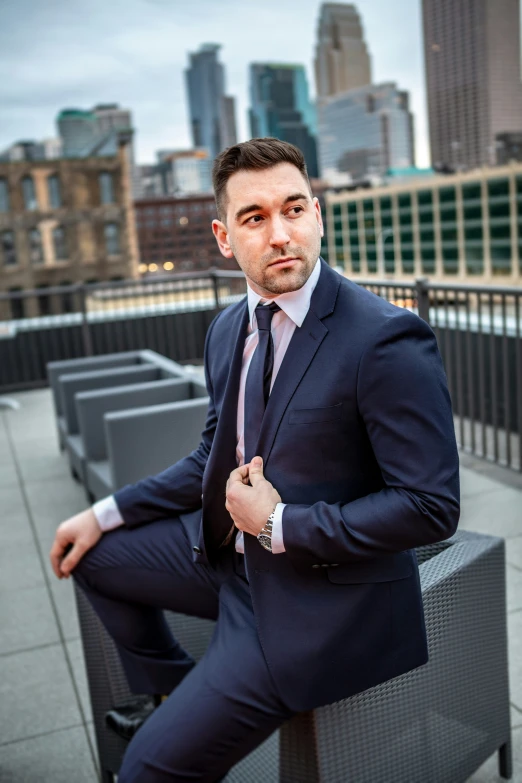 man in a suit is sitting on a bench with the city behind him