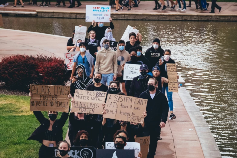 group of people standing next to a body of water holding placards and wearing face masks