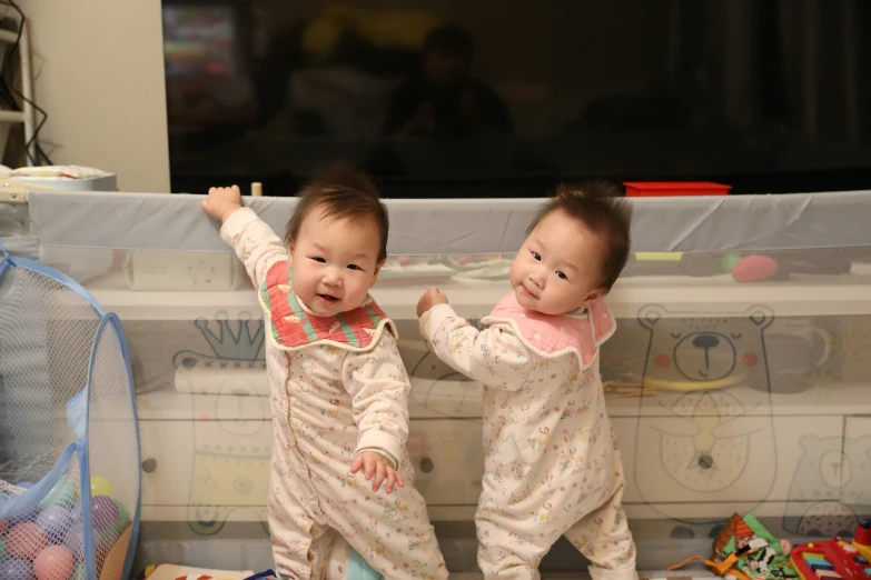 two babies in pajamas standing up against a wall