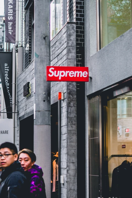 two people walk past some shops with a sign saying supreme