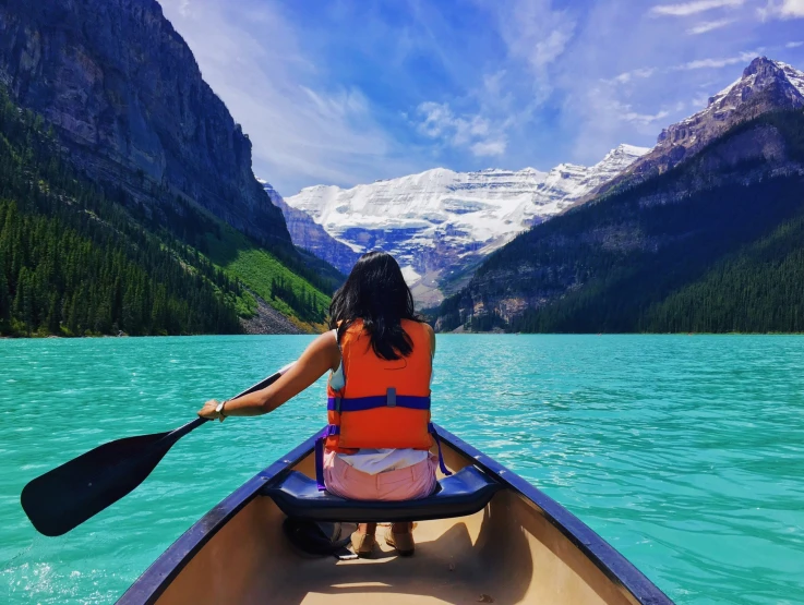 a woman in an orange life jacket rows her canoe down a mountain lake