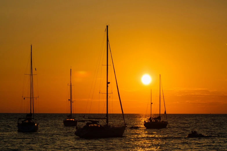 the setting sun with sailboats in the ocean and people on beach