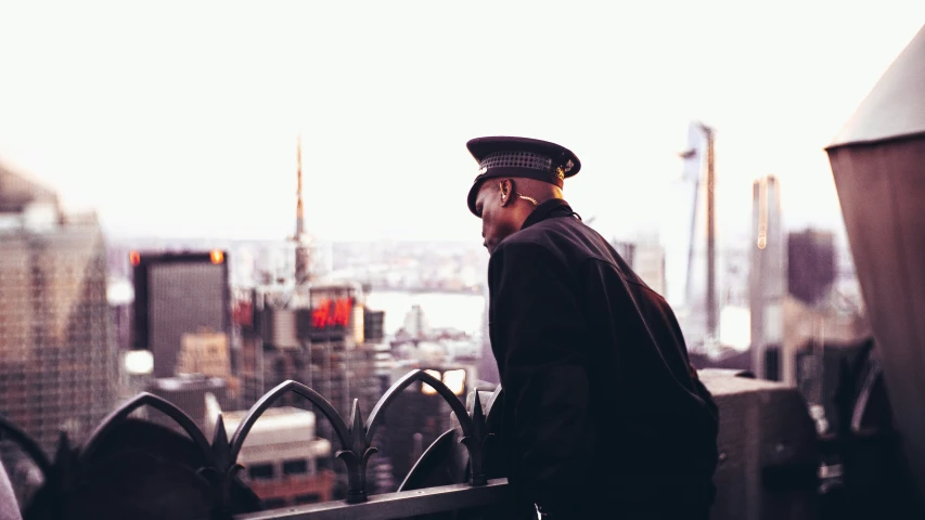 a man in hat standing on a roof top with view of an urban city