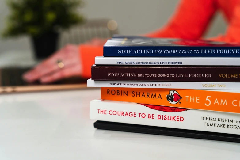 a stack of books on a table with a red vase and orange chair in the background