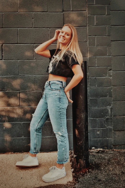 a young blond woman in boyfriend jeans leaning against a brick wall