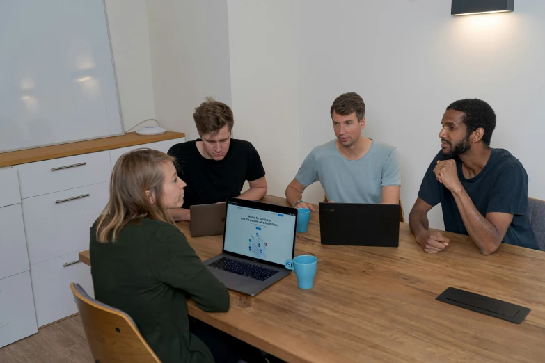 four people sitting at a table with laptops and talking to each other