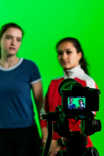 two young women are standing in front of green screen while the third person uses a camera