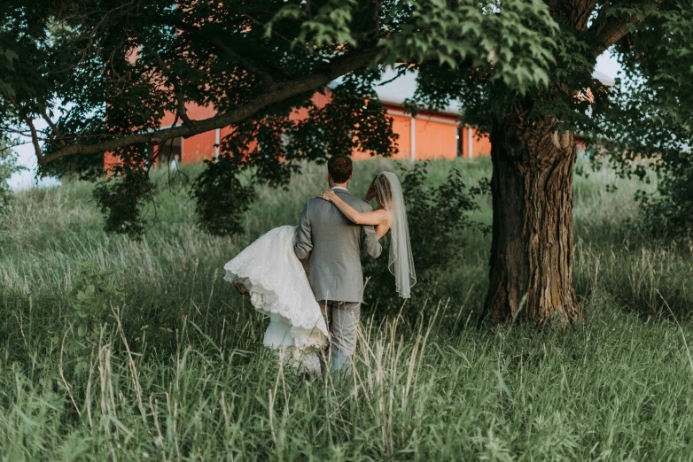 a man and woman walking through tall grass near trees