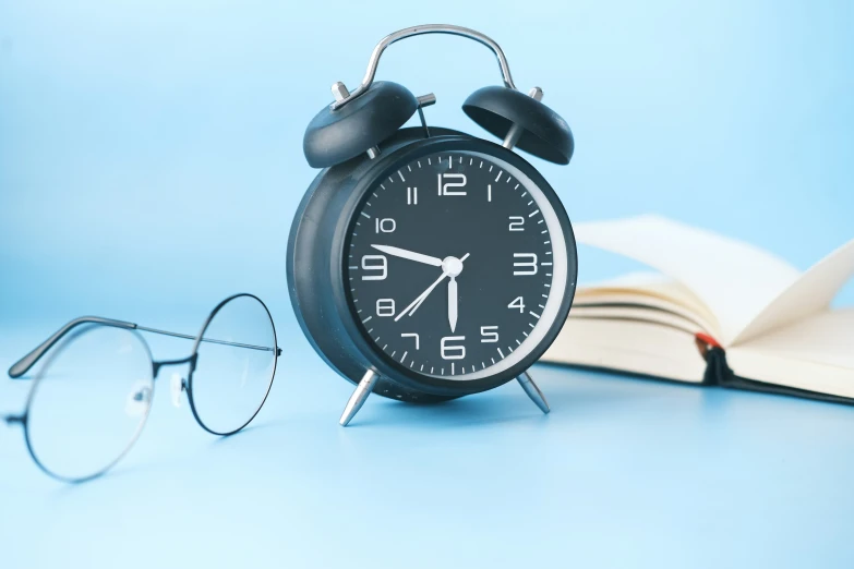 an alarm clock next to a book, eyeglasses, and pen