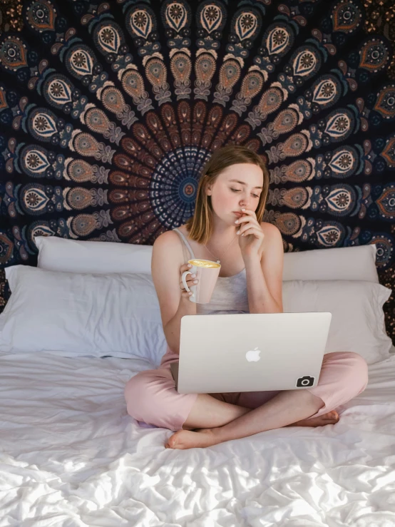 a girl sitting on a bed with a laptop and beverage in hand