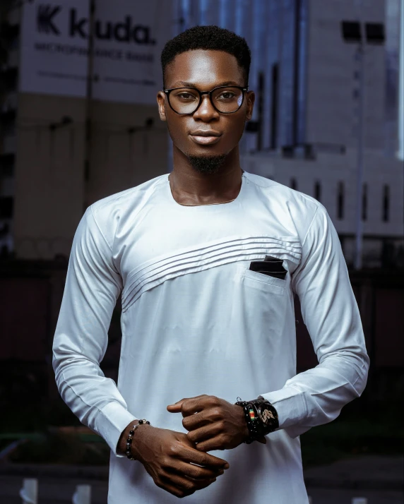 a man with glasses wearing white t - shirt, standing in front of buildings