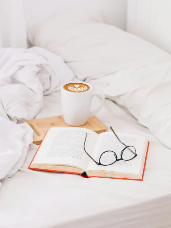 a cup of coffee, eyeglasses, and an open book on a bed