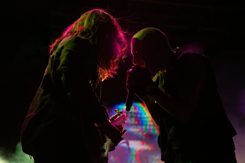 two people on stage during a concert with the light from brightly colored lights