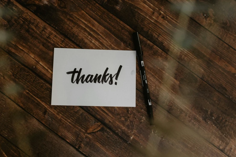 a piece of paper that says thanks on a wooden surface with some pens next to it