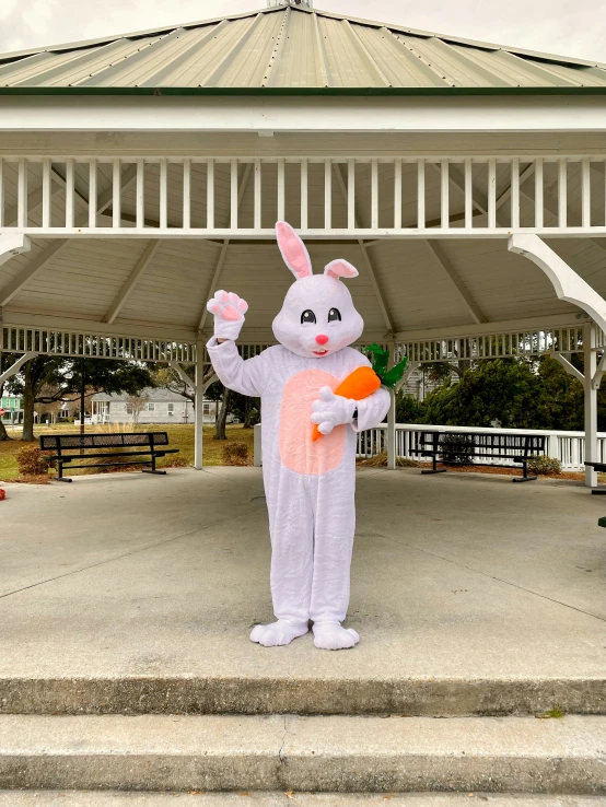 the man in an adult easter bunny suit stands near a gazebo and holds up a carrot