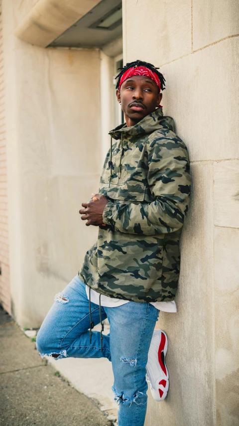 a man leans against a building wall wearing a camo jacket