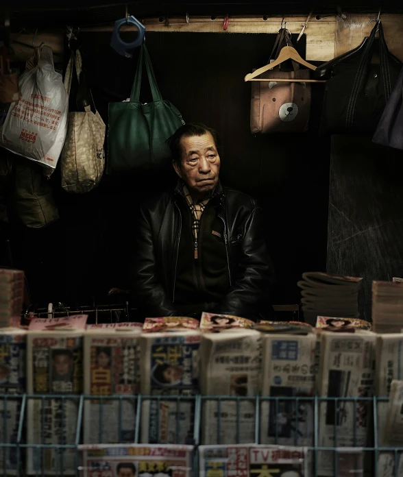 man in black jacket sitting under newspaper stand with multiple bags on it