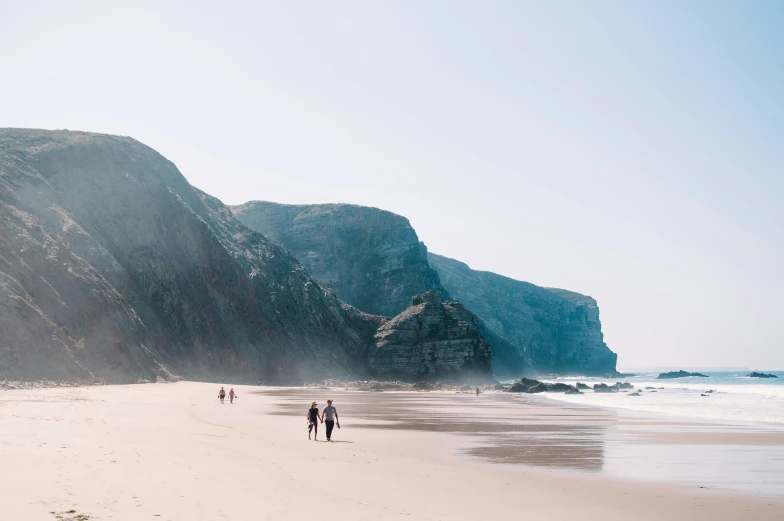 three people are walking along the beach near a cliff