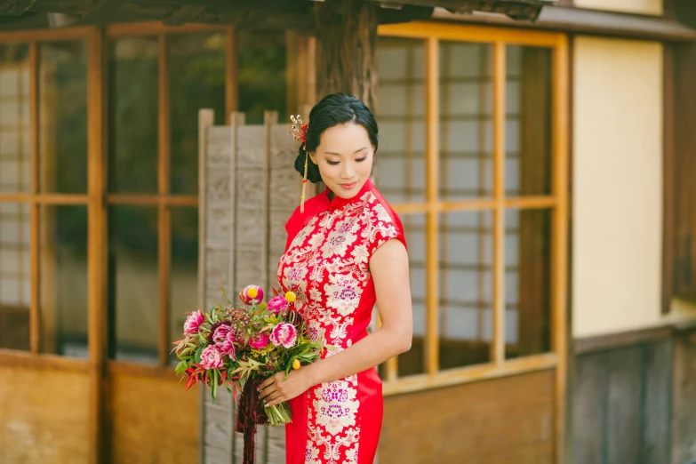 a woman in red holding flowers standing outside a hut