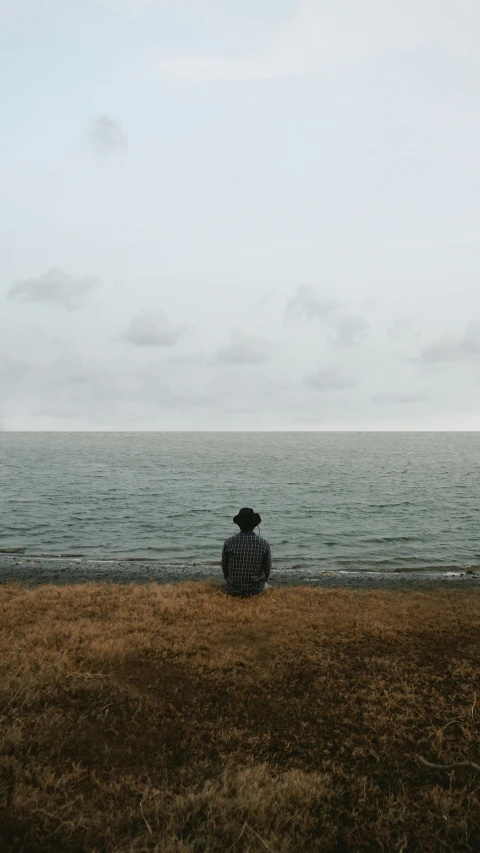 the man is sitting on the beach facing the water