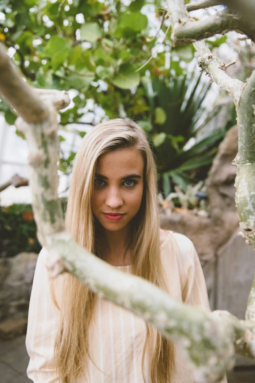 an attractive woman in a white top is standing near a tree