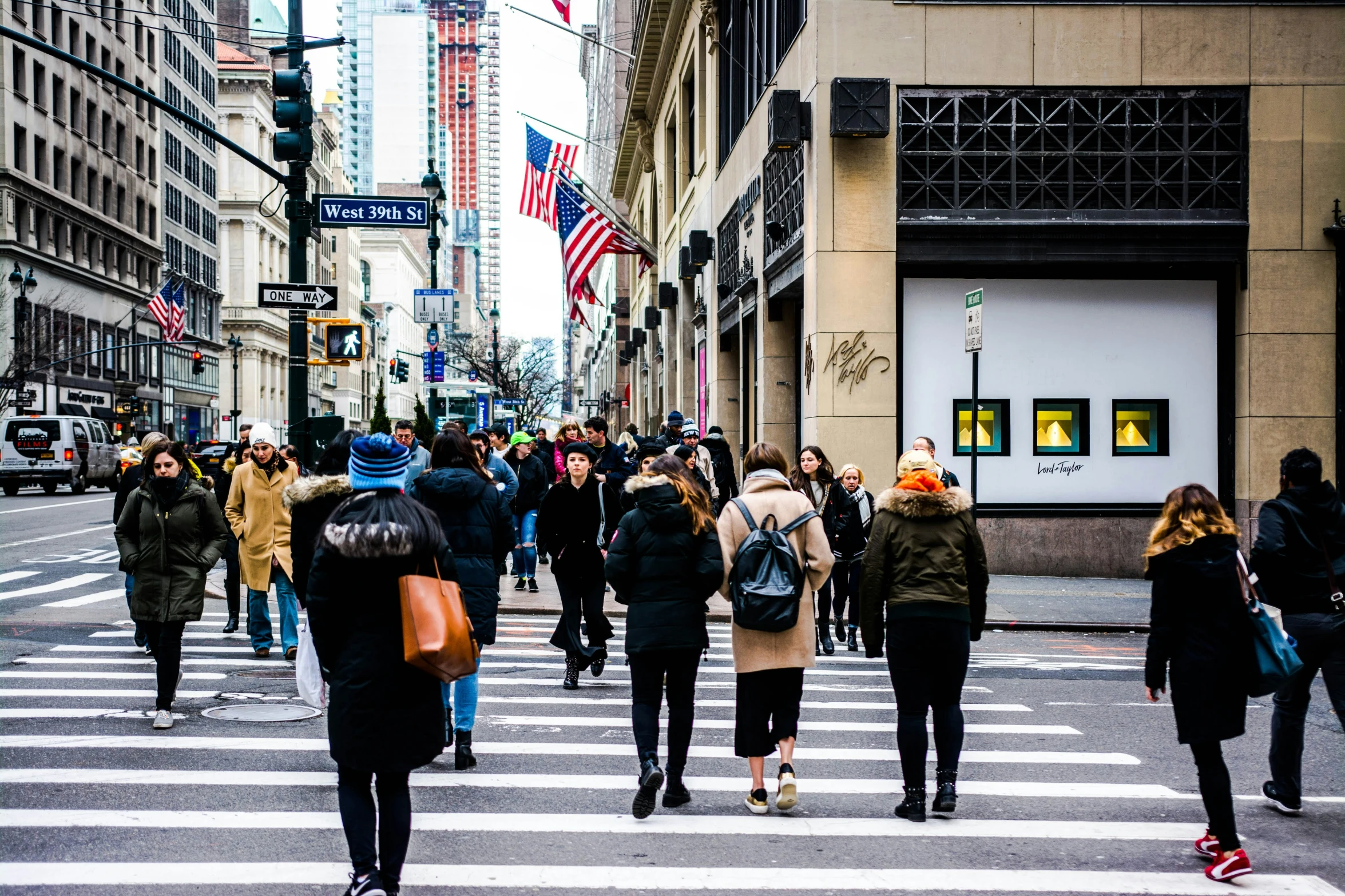 people walk across an urban street with a lot of flags on the building and lights