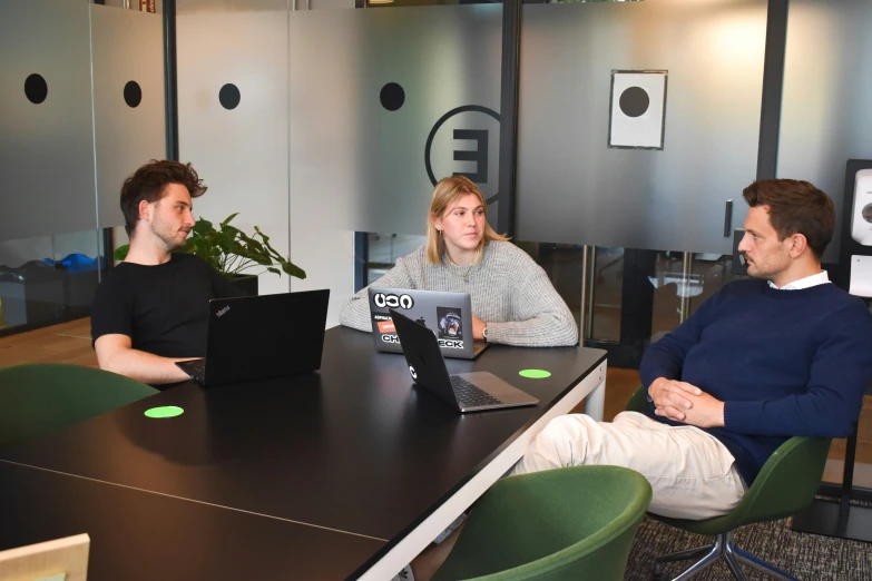 three people are sitting at a long table with two of them working on laptops