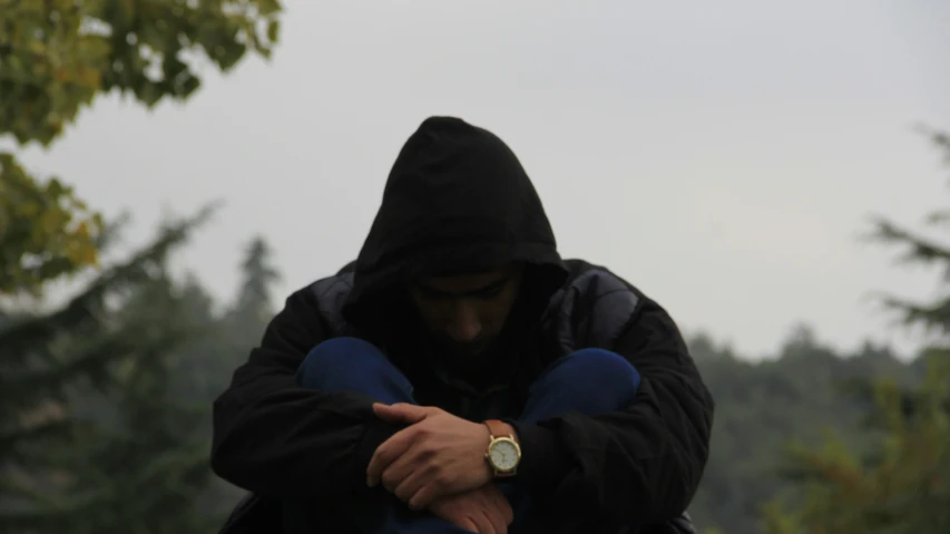 a man is covering his face with one hand while standing on a bench in front of some trees