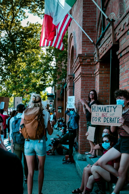 a woman holds up a sign while others sit on the curb in front of a brick building