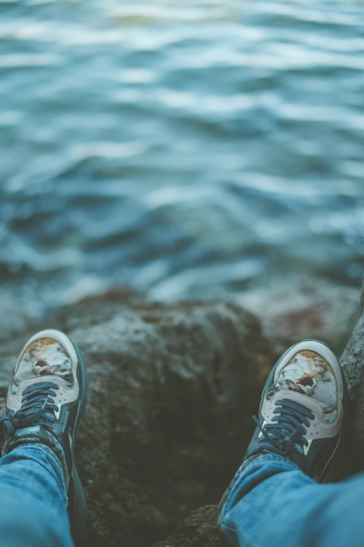 a person wearing blue jeans and white sneakers is seen standing on some rocks near the water