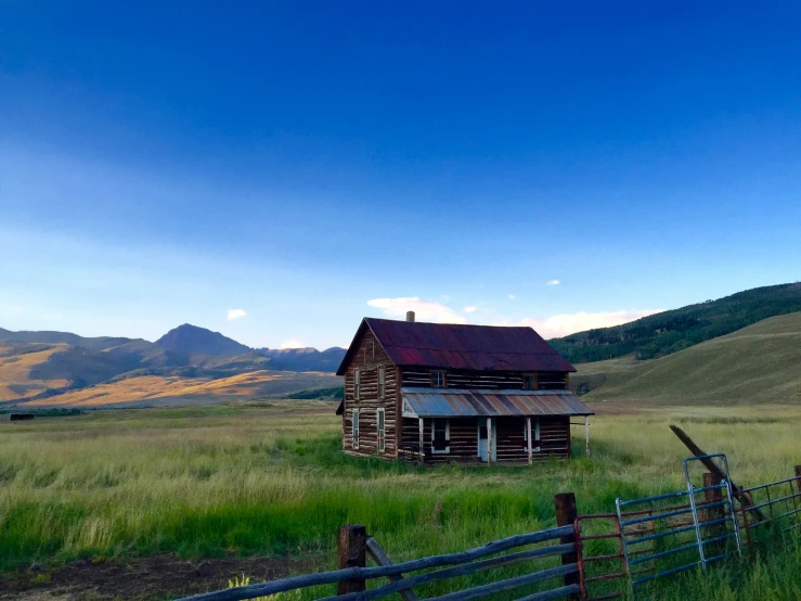 a rustic log cabin stands in the middle of a large open field with mountains and hills behind it